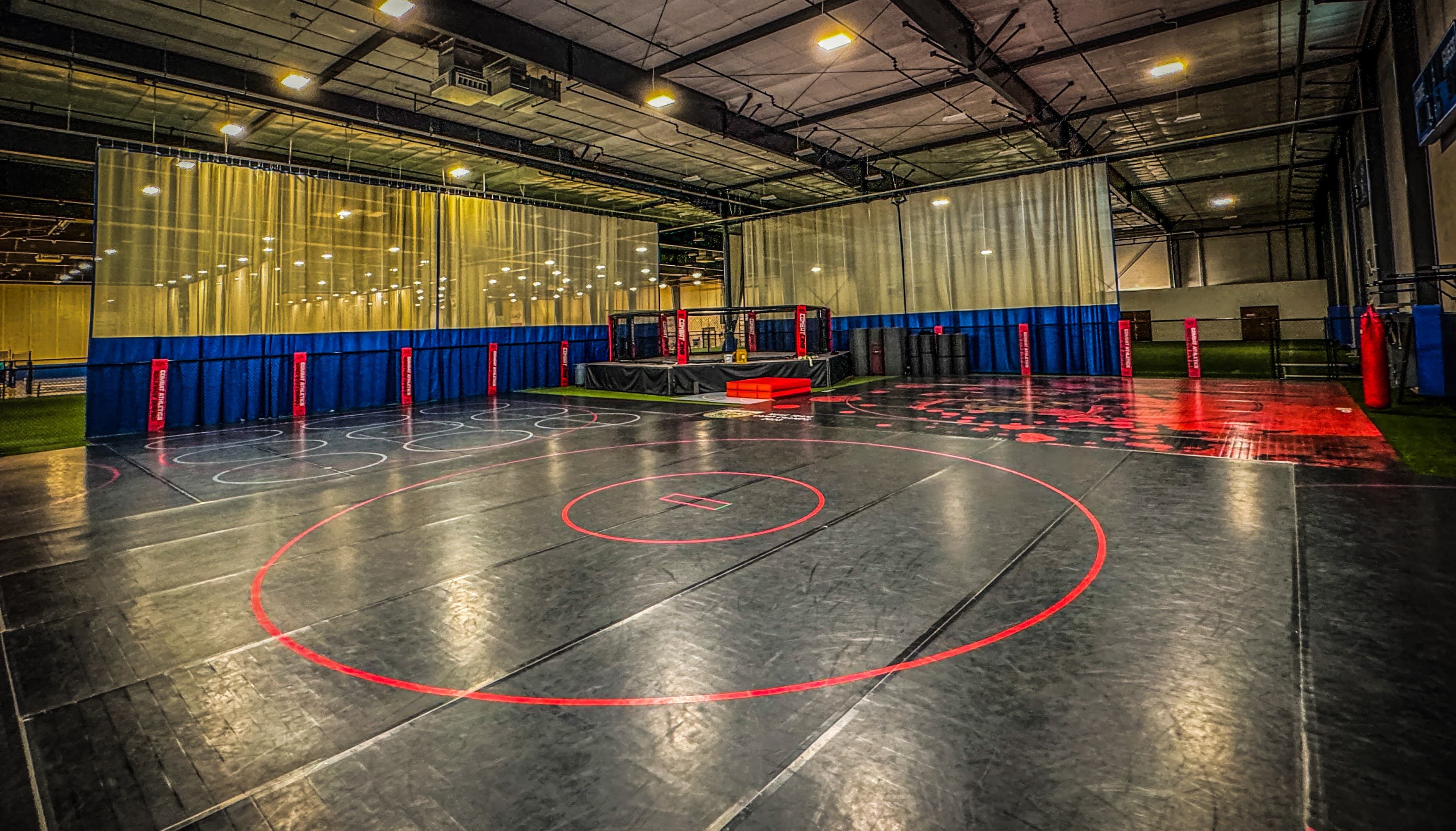 The Combat Athletics training floor: black wrestling mats with red circles and a fight cage